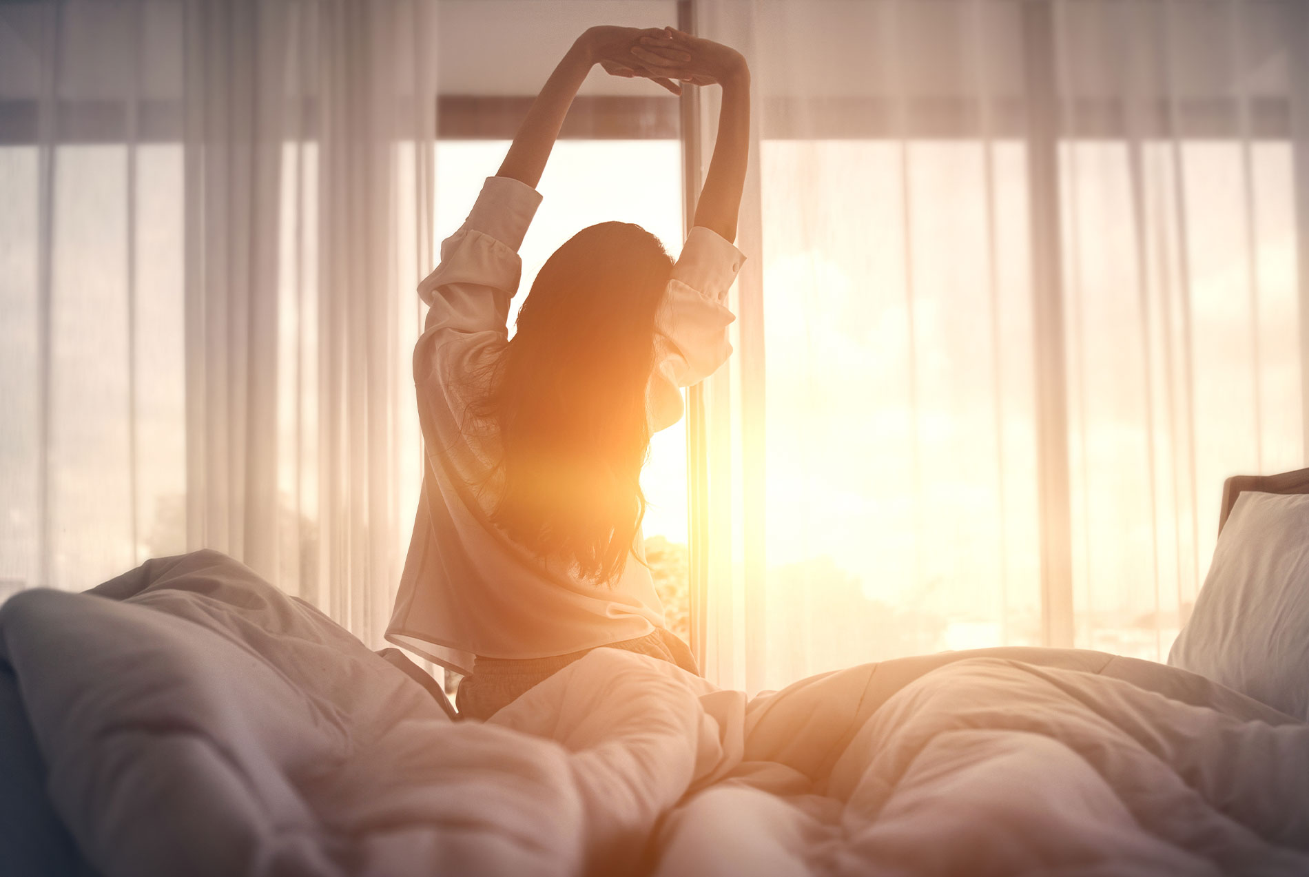 A person is stretching their arms above their head while lying on a bed with sunlight streaming through a window.